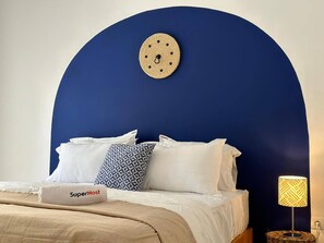 Un dormitorio con cabecera azul, ropa de cama blanca, almohadas estampadas y una lámpara de noche.