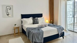Ein modernes Schlafzimmer mit dunkelblauem Kopfteil, einem Nachttisch, einer kleinen Topfpflanze und Blick auf einen Balkon mit Geländer.