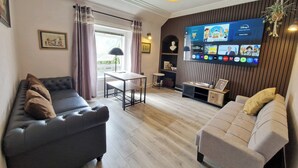 Un salon moderne comprenant un canapé, une petite banquette, une table et un téléviseur fixé au mur, qui diffuse différentes chaînes.