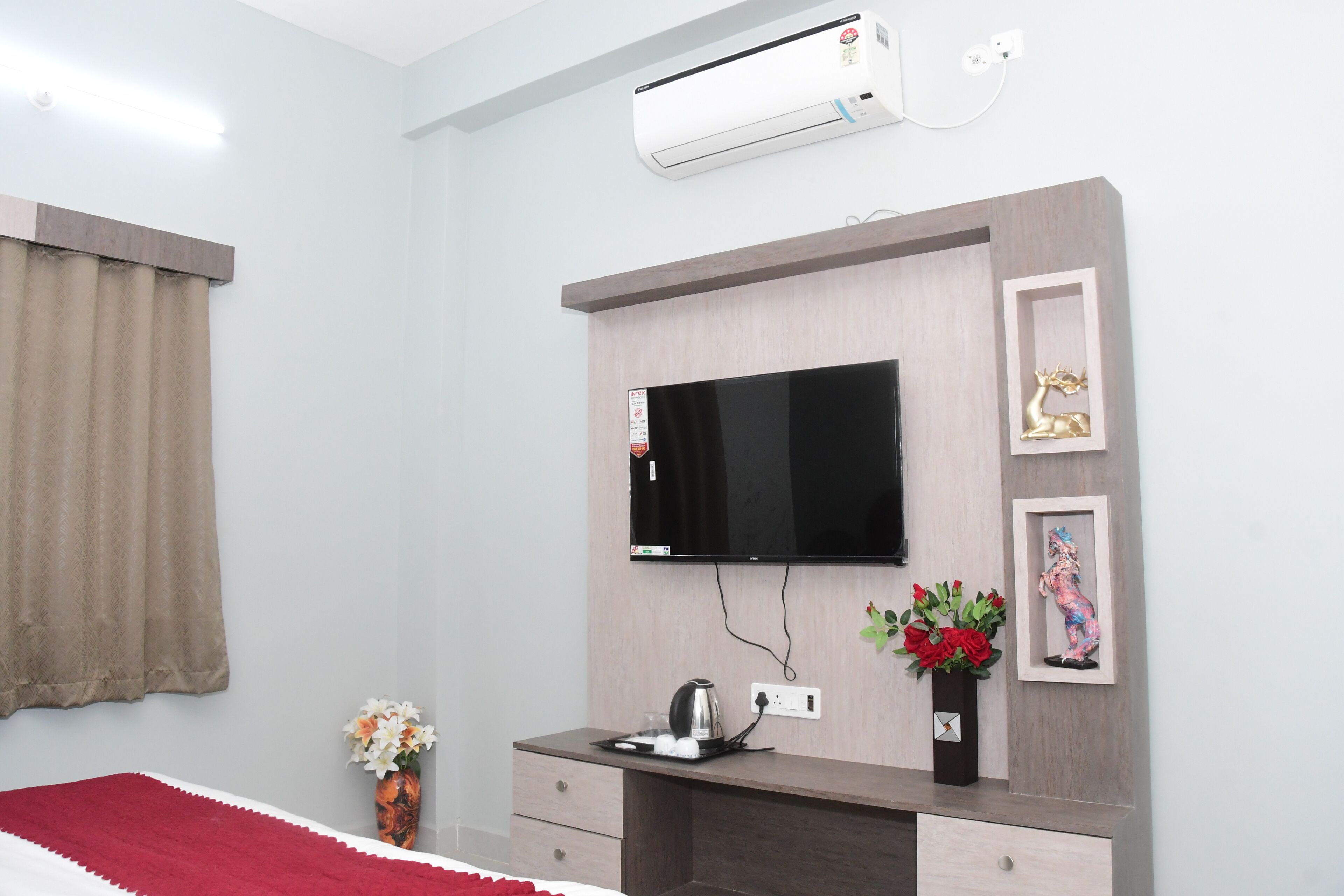 A hotel room with a bed, a TV mounted on the wall, a desk with a kettle, and a vase with flowers.