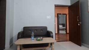 A modern room with a black leather sofa, a wooden table with a bottle and bowl, a wooden door, and a hallway leading to a bed.