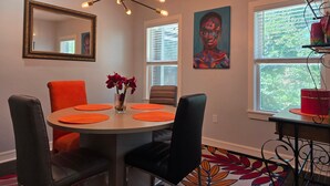 A dining area with a round table, orange placemats, black chairs, a vase with red flowers, a modern chandelier, a framed mirror, a colorful painting, and a window with blinds. - World Cup Ready - Near Downtown & Museum District (Houston)