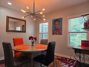 A dining area with a round table, orange placemats, black chairs, a vase with red flowers, a modern chandelier, a framed mirror, a colorful painting, and a window with blinds. - World Cup Ready - Near Downtown & Museum District (Houston)