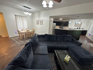 Ein Wohnzimmer mit einem dunkelgrauen Ecksofa, einem Glastisch und einem Essbereich mit Holzsesseln und einem Tisch.