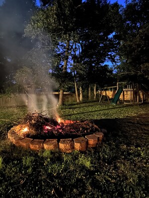 Ein Lagerfeuer in einer runden Steinkaminkasse, umgeben von Gras, mit Bäumen und einer Holzhütte im Hintergrund.