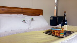 A neatly made bed with a yellow blanket, two pillows with a logo, a bottle of La Pluma, and a tin of food on a tray.