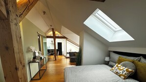 Ein Schlafzimmer mit Oberlichter, Holzbalken, einem Bett mit gemusterten Betttextilien und Blick auf eine Küche und einen Essbereich.