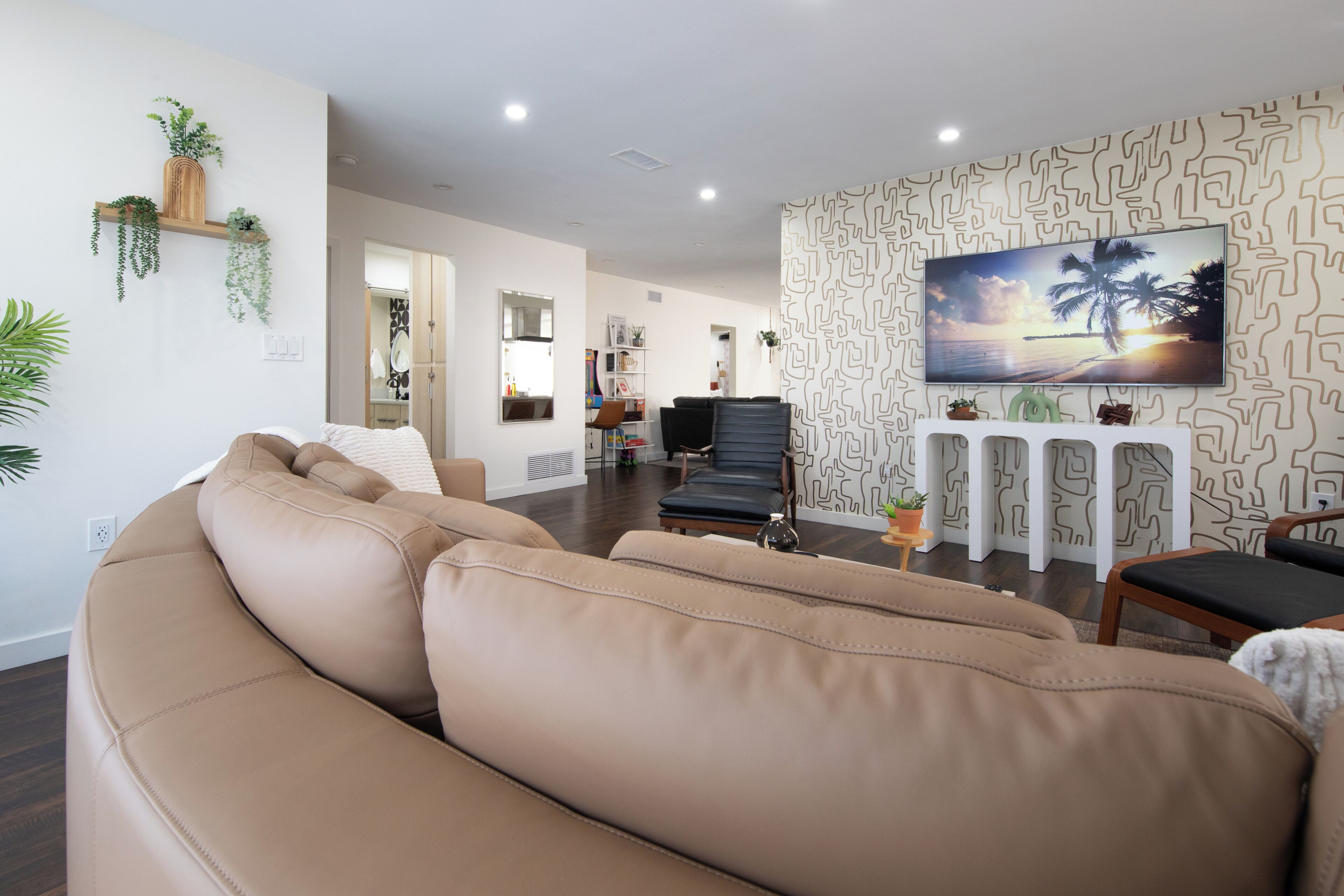 Een woonkamer met een beige bank, een flatscreen televisie aan een patroonmuur, en een houten plank met planten.