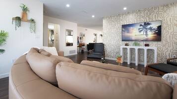 Een woonkamer met een beige bank, een flatscreen televisie aan een patroonmuur, en een houten plank met planten.