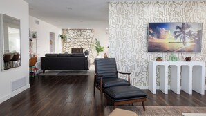Ett modernt vardagsrum med en vägg som har en dekorativ stenbeklädnad, en svart lädersoffa, en svart läderfåtölj, ett vitt konsolbord med dekorativa föremål och en stor tavla som föreställer en solnedgång över havet.