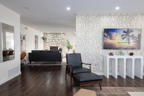 Ein modernes Wohnzimmer mit einer Wand aus Stein, einem schwarzen Ledersofa, einem schwarzen Lederstuhl, einem weißen Konsolentisch mit Dekoration und einem großen Gemälde, das einen Sonnenuntergang über dem Meer zeigt.