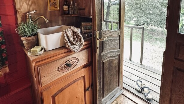 A wooden cabin interior with a wooden counter, a sink, a door leading outside, and a view of the outdoors.
