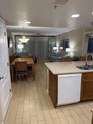 Eine kompakte Küche mit Geschirrspüler, ein Essbereich mit Stühlen und Tisch sowie ein Wohnzimmer mit Sofa und Vorhängen.