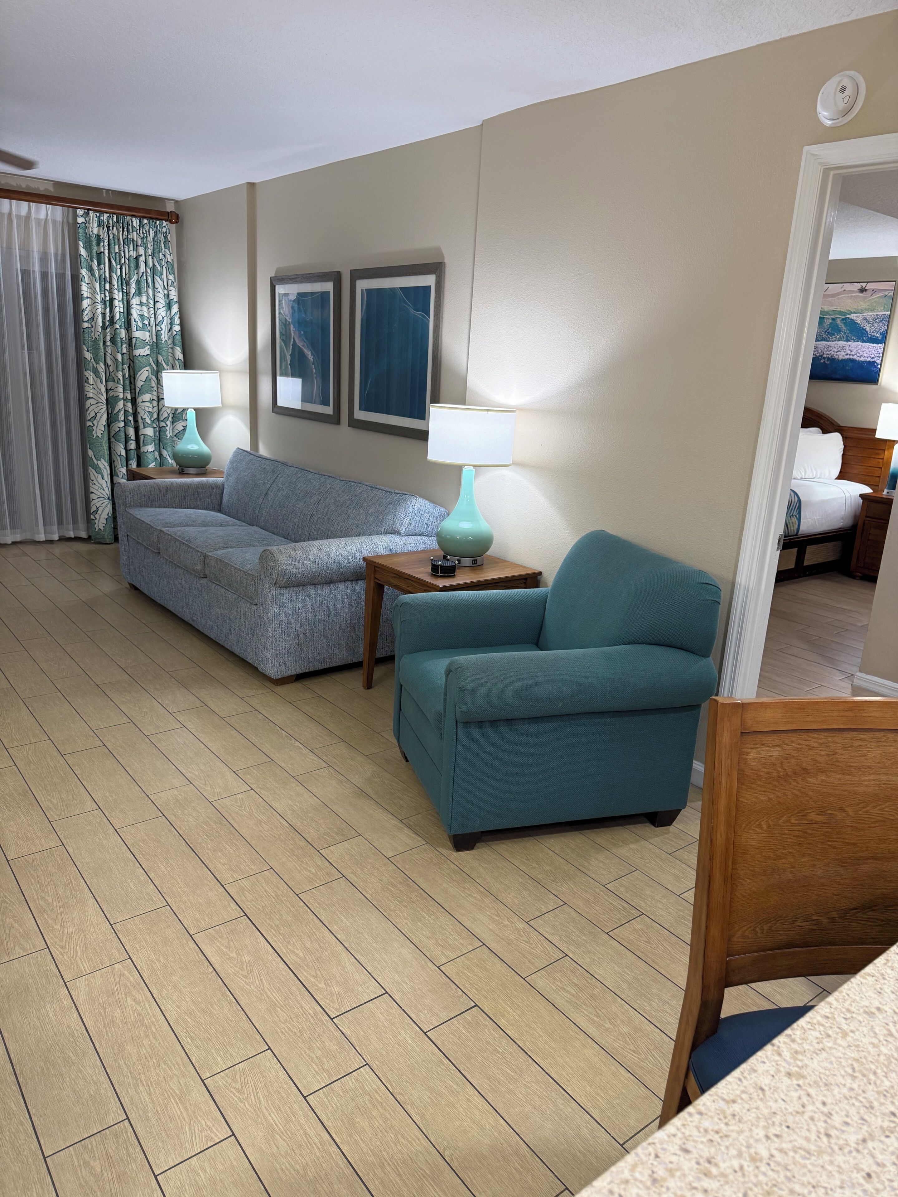 Una habitación de hotel con un sofá gris, un sillón verde azulado, una mesita de madera pequeña con una lámpara verde azulado y una cama con ropa blanca.