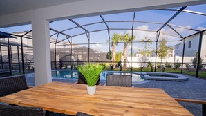 A wooden table with wicker chairs and a potted plant in a covered outdoor area with a view of a swimming pool and palm trees. - Brand New 10BR 8BA/Themed BR/PrivatePool/SPA 16276 (Clermont)