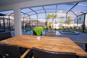 Ein Holztisch mit Korbstühlen, einem Blumentopf und einer gläsernen Veranda mit Pool und Palmen.