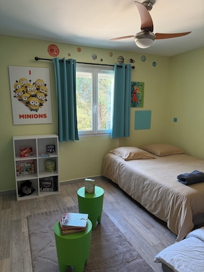 A bedroom with a bed, a window with curtains, a ceiling fan, a bookshelf with board games, and a green stool.