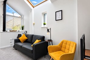 Een moderne woonkamer met een zwarte bank, een gele fauteuil, een bijzettafeltje en een gecertificeerd diploma aan de muur.