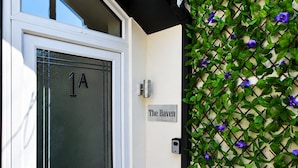 Una puerta con una placa de número, un letrero en la pared y una celosía con follaje verde y flores púrpuras.