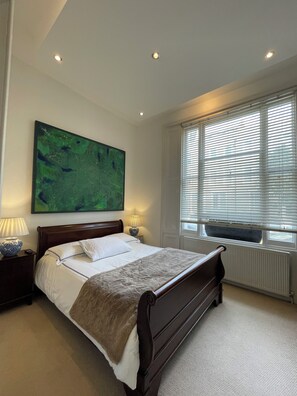 Ein Schlafzimmer mit einem Bett, einem Nachttisch, einer Lampe, einem Gemälde und einem Fenster mit Jalousien.