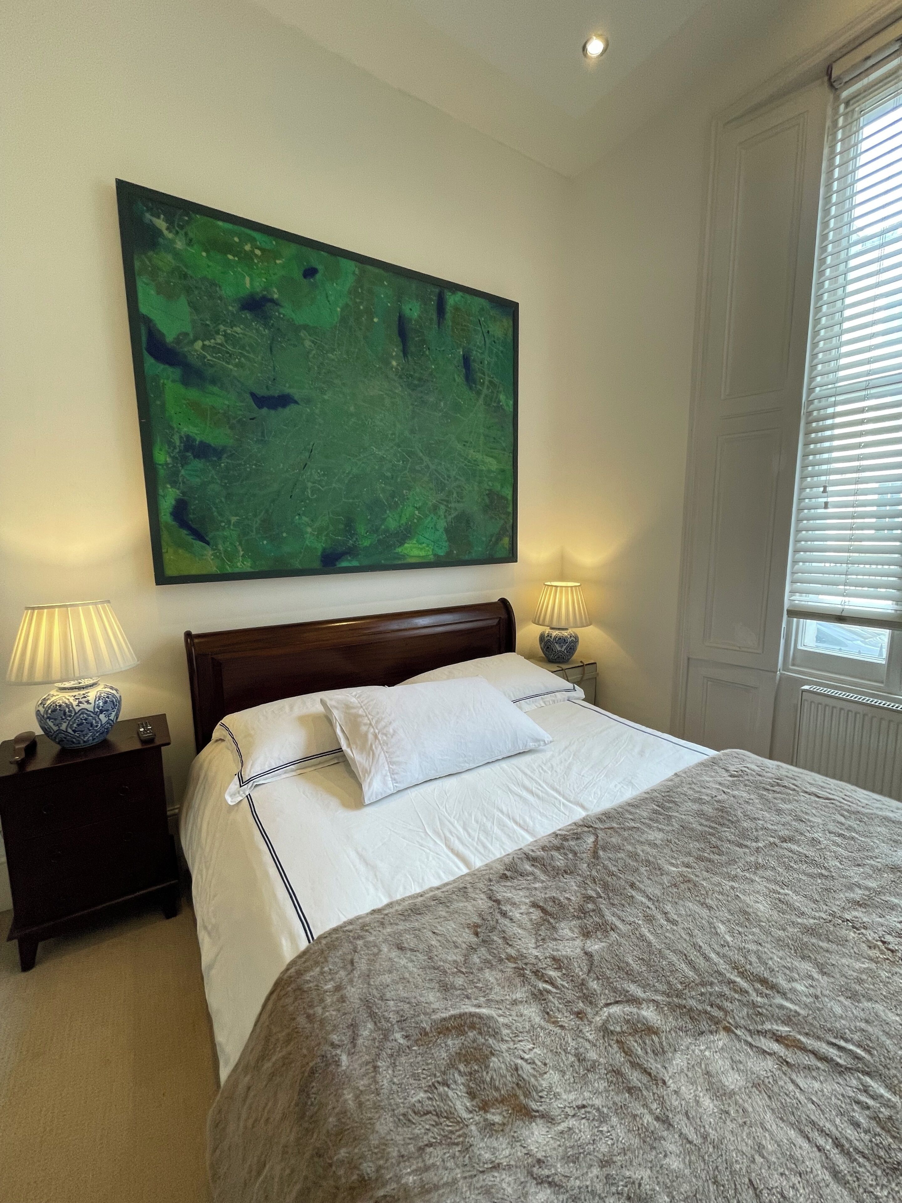 A bedroom with a large bed, two bedside tables, a nightstand, a green abstract painting, and a window with blinds.