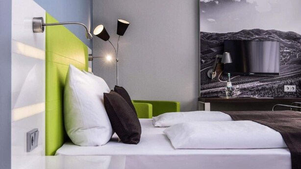 A modern hotel room with a bed, green headboard, white bedding, a wall-mounted TV, and a landscape painting. - 1 Person Holiday Park Home in Offenburg (Offenburg)