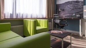 A modern hotel room with a green sofa, a small table, a chair, a radiator, and a large window with sheer curtains. - 1 Person Holiday Park Home in Offenburg (Offenburg)