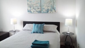 A neatly made bed with white and blue pillows, two bedside lamps, and a pair of wall art depicting flowers. - Cozy Fully Furnished Apt w/ Parking (Toronto)