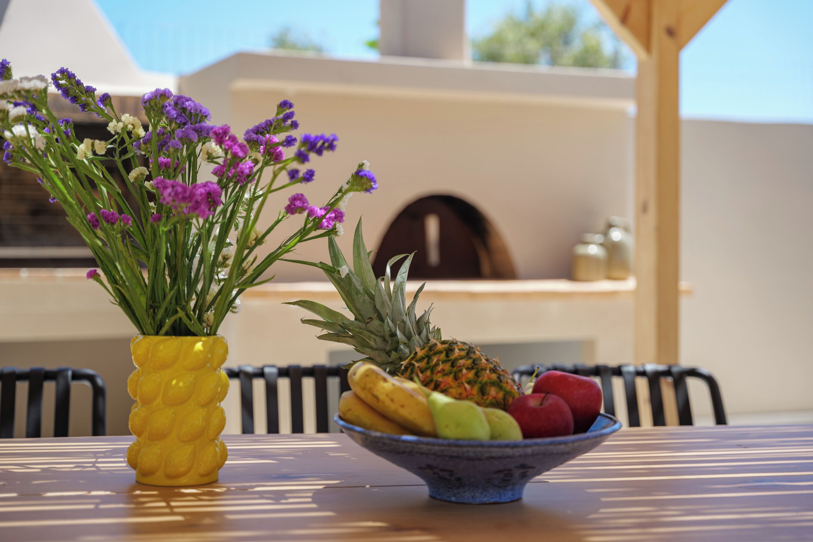 A table with a yellow vase of purple flowers and a bowl of fruit.
