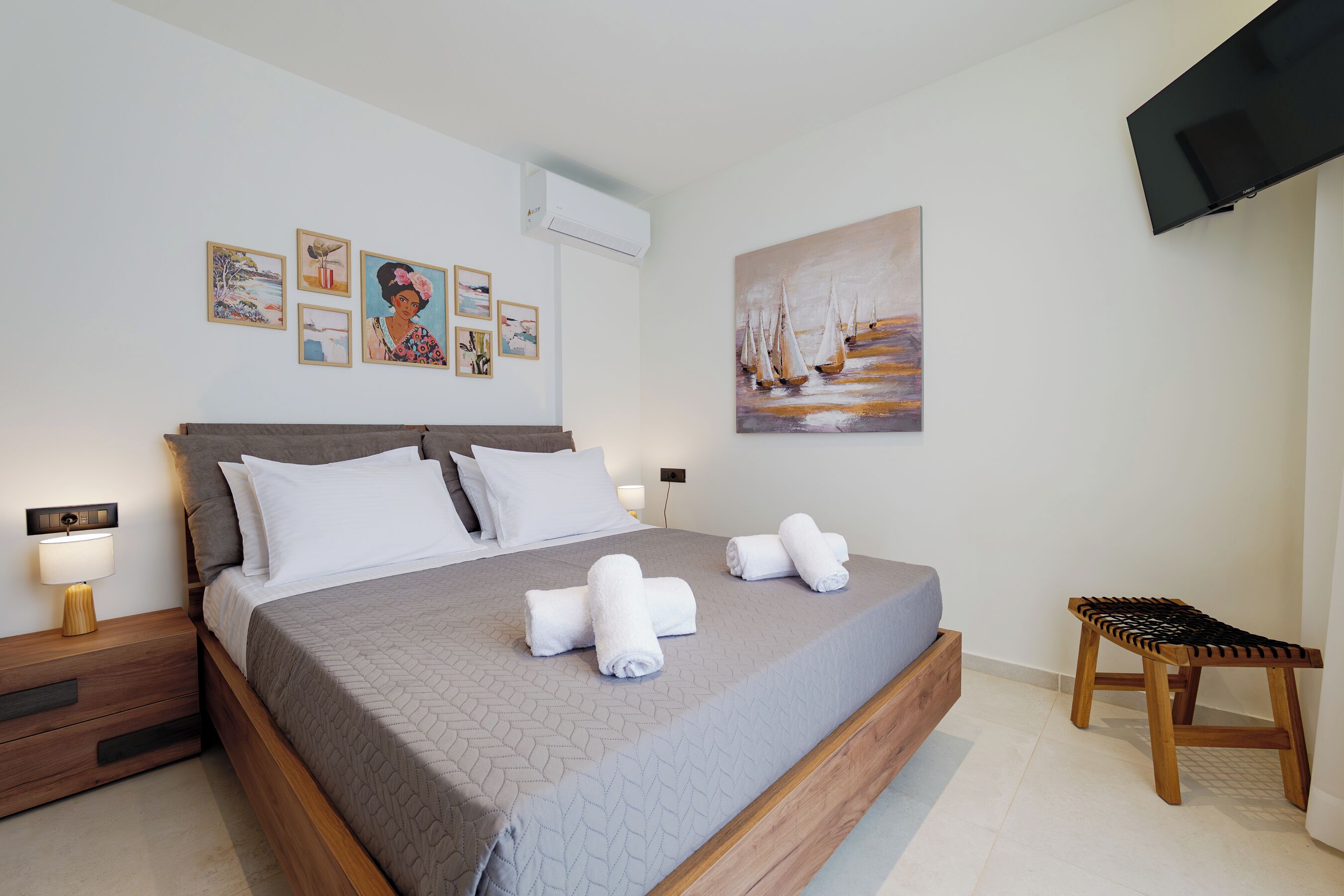 A neatly made bed with white linens, a wooden nightstand with a lamp, and a painting of sailboats on the wall.