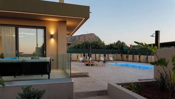 A modern outdoor pool area with lounge chairs, a glass balustrade, and a view of a mountain. - Verde De Luna Villa (Hersonissos)