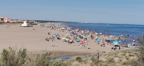 Ein belebter Strand mit badenden und sonnenden Menschen, einem Rettungsturm und einem wolkenlosen Himmel.