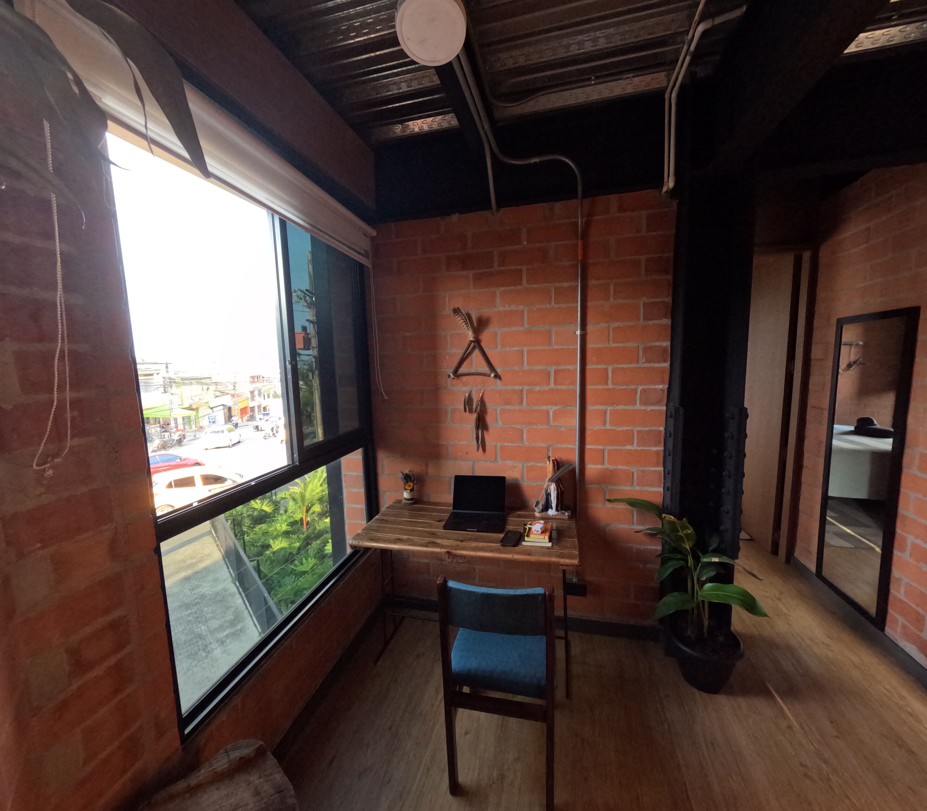 A room with a brick wall, a wooden desk, a chair, a laptop, and a large window.