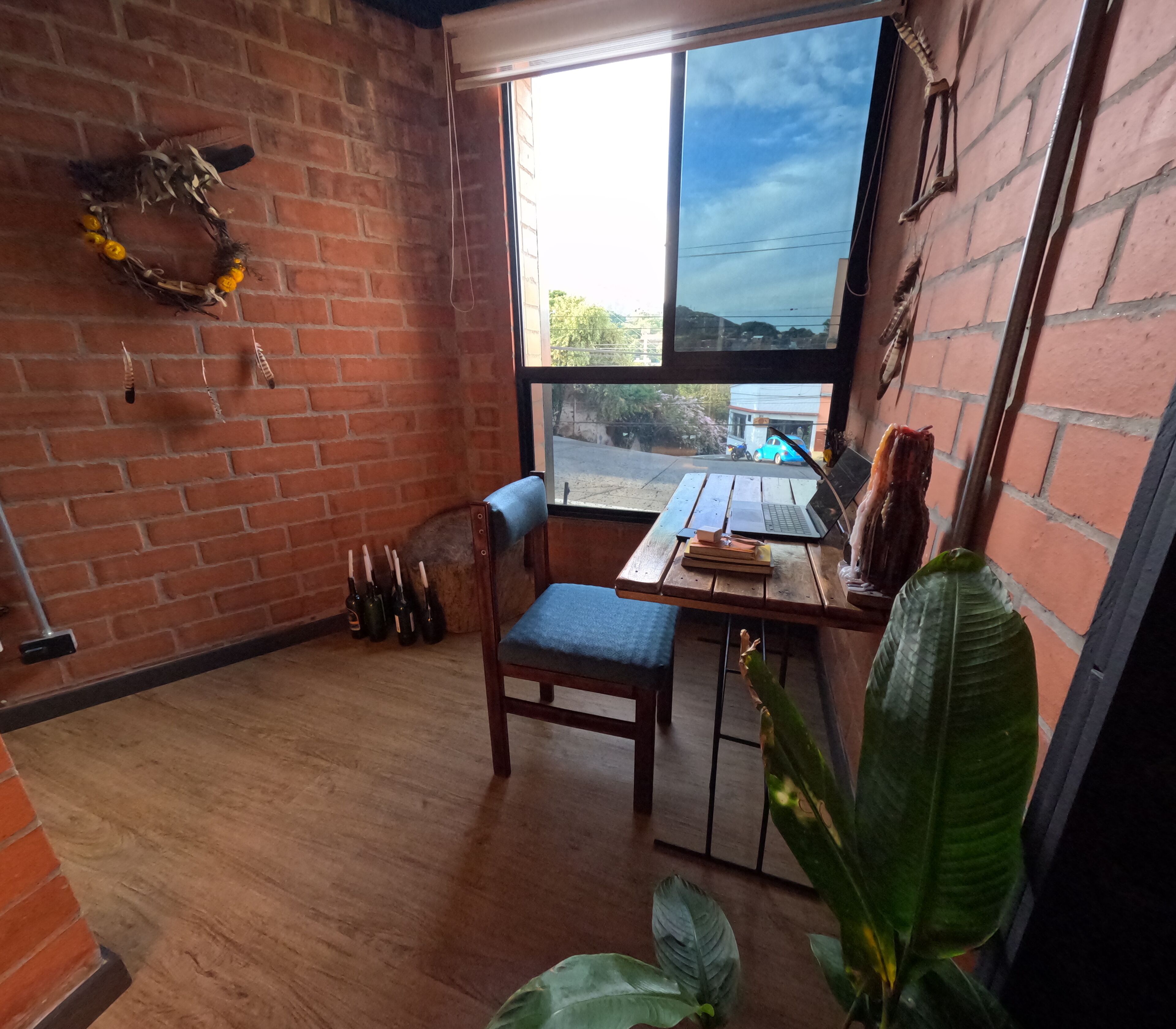 A room with a brick wall, a wooden floor, a window with a view of the street, a table with a laptop, a chair, and a plant.