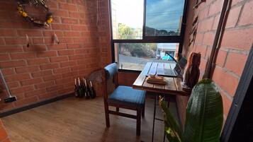 A room with a brick wall, a wooden floor, a window with a view of the street, a table with a laptop, a chair, and a plant.