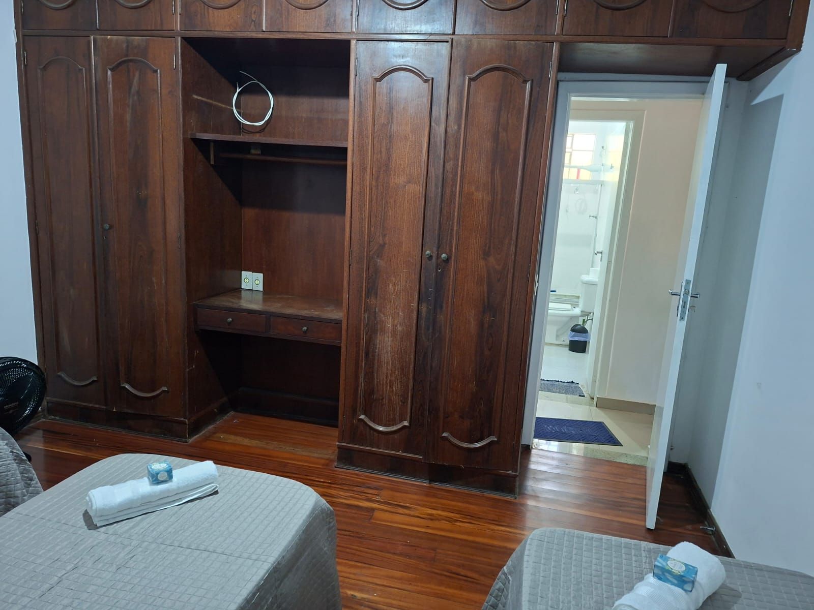 A bedroom with a wooden wardrobe, a bed with a grey bedspread, and a small bedside table with a bottle and a tissue box.