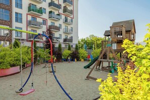 Ein Spielplatz mit Schaukeln, Rutschen und Klettergeräten, umgeben von einem Gebäude mit Balkonen und einem hölzernen Spielhäuschen.