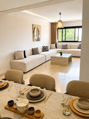 A modern living room with a sofa, dining table, and a large window. - Luxury 3BR Apt - Gated Residence - Pools - City Center (Marrakech)