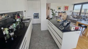 Eine moderne Küche mit weißen Schränken, einer schwarzen Arbeitsplatte und einem eingebauten Backofen. Ein Essbereich mit einem Holztisch und Stühlen bietet einen Blick auf die Stadt durch ein großes Fenster.