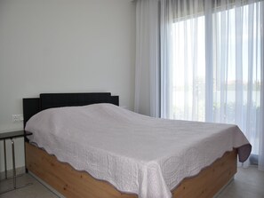 Ein Einzelbett mit Holzrahmen und weißer Bettdecke in einem Zimmer mit großem Fenster und durchsichtigen Vorhängen.