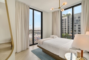 Ein Hotelzimmer mit Bett, Nachttisch, Lampe und großen Fenstern mit Blick auf eine Stadtlandschaft.