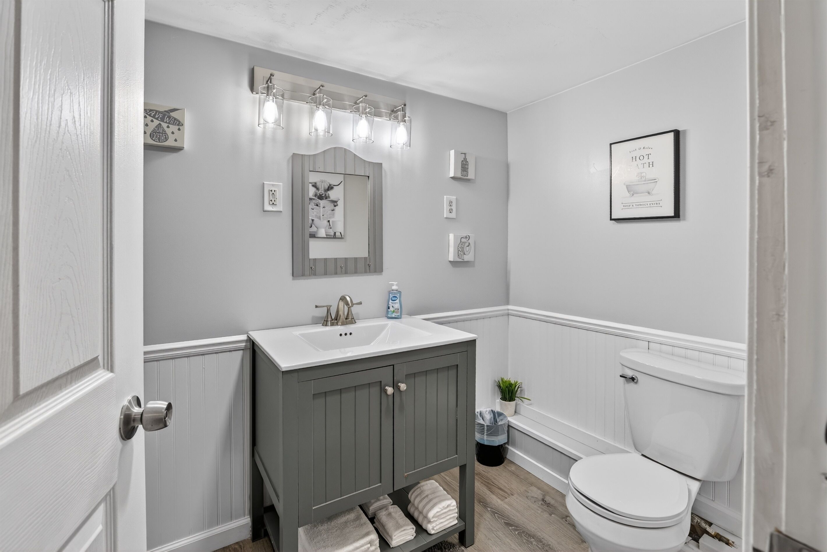 Un baño con lavamanos blanco, tocador gris, inodoro blanco y espejo con luces arriba.