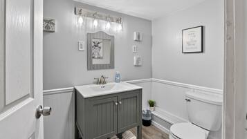 Un baño con lavamanos blanco, tocador gris, inodoro blanco y espejo con luces arriba.