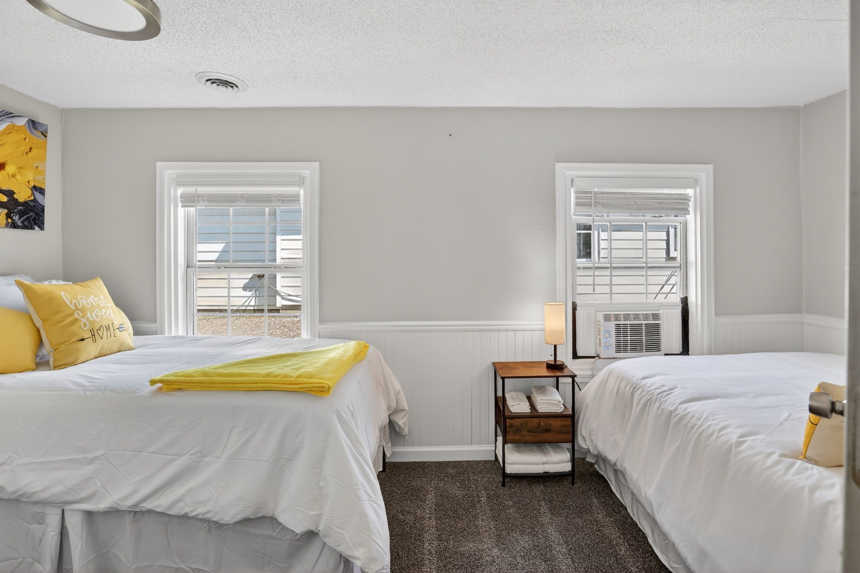 A hotel room with two beds, a small table with a lamp, a radiator, and a window with a view.
