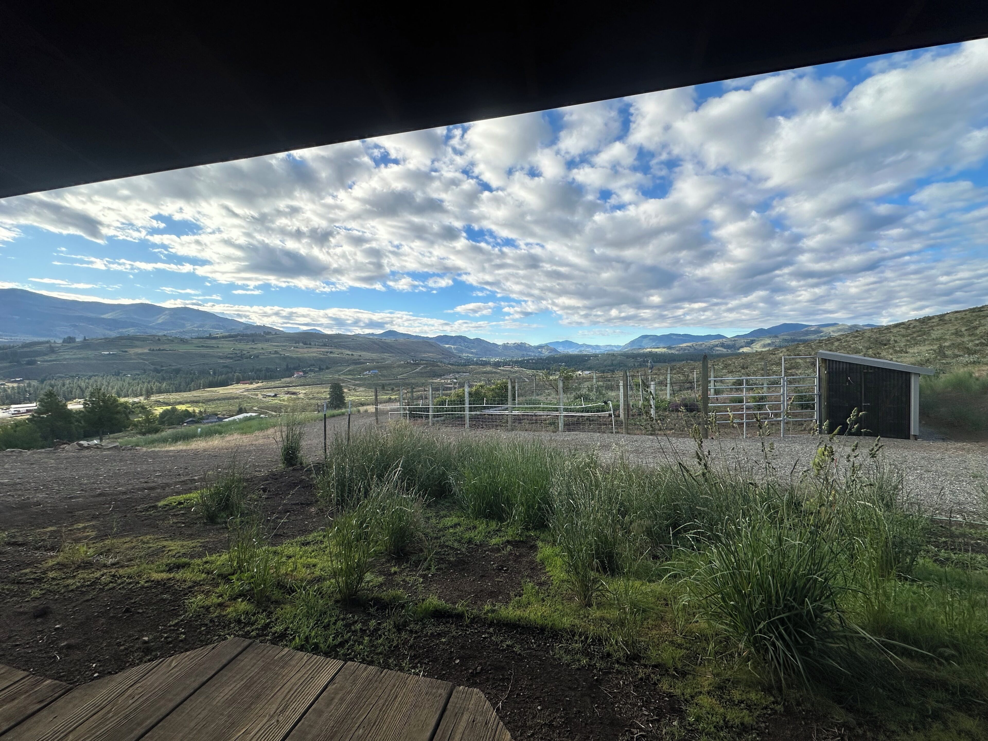 Un paysage rural avec une zone délimitée par une clôture, un petit bâtiment et des montagnes à l’horizon.
