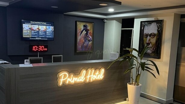 Ein Empfangsschalter mit einem Neonzeichen, das 'Primal Hotel' zeigt, einer digitalen Uhr, auf der 22:30 Uhr angezeigt wird, und einem Fernsehbildschirm darüber.