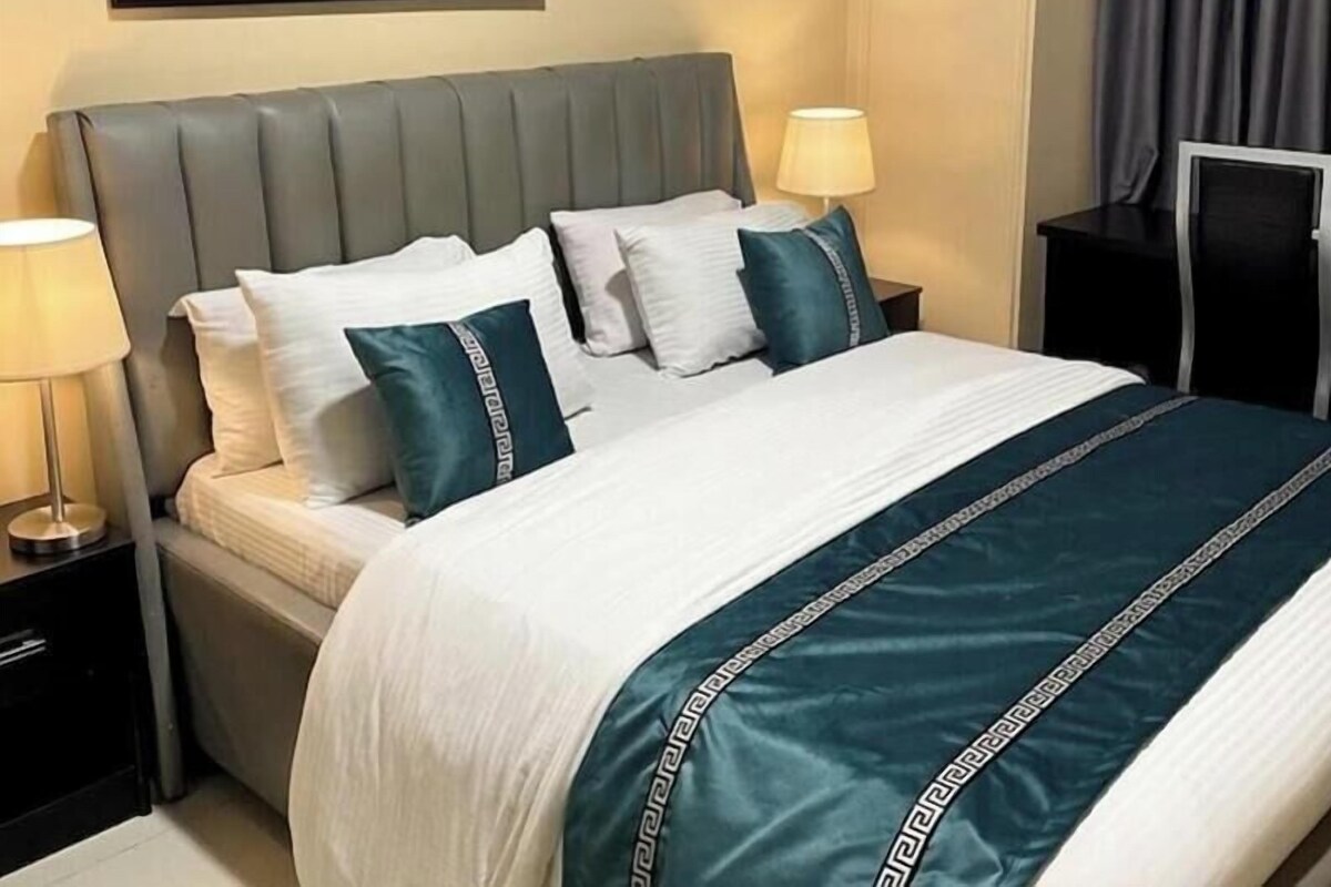 Habitación de hotel con cama, almohadas, cabecero, mesita de noche, lámpara, un cuadro en la pared y una ventana con cortinas.