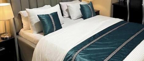 Habitación de hotel con cama, almohadas, cabecero, mesita de noche, lámpara, un cuadro en la pared y una ventana con cortinas.
