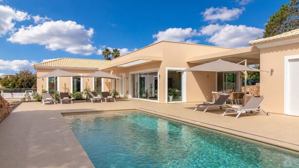 Ein moderner Poolbereich mit Liegestühlen und Sonnenschirmen, einem kühlen Pool und einem Gebäude mit großen Fenstern und einer flachen Dachkonstruktion.