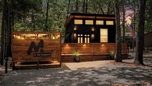 A small, modern wooden cabin with a large letter 'M' sign, surrounded by trees.